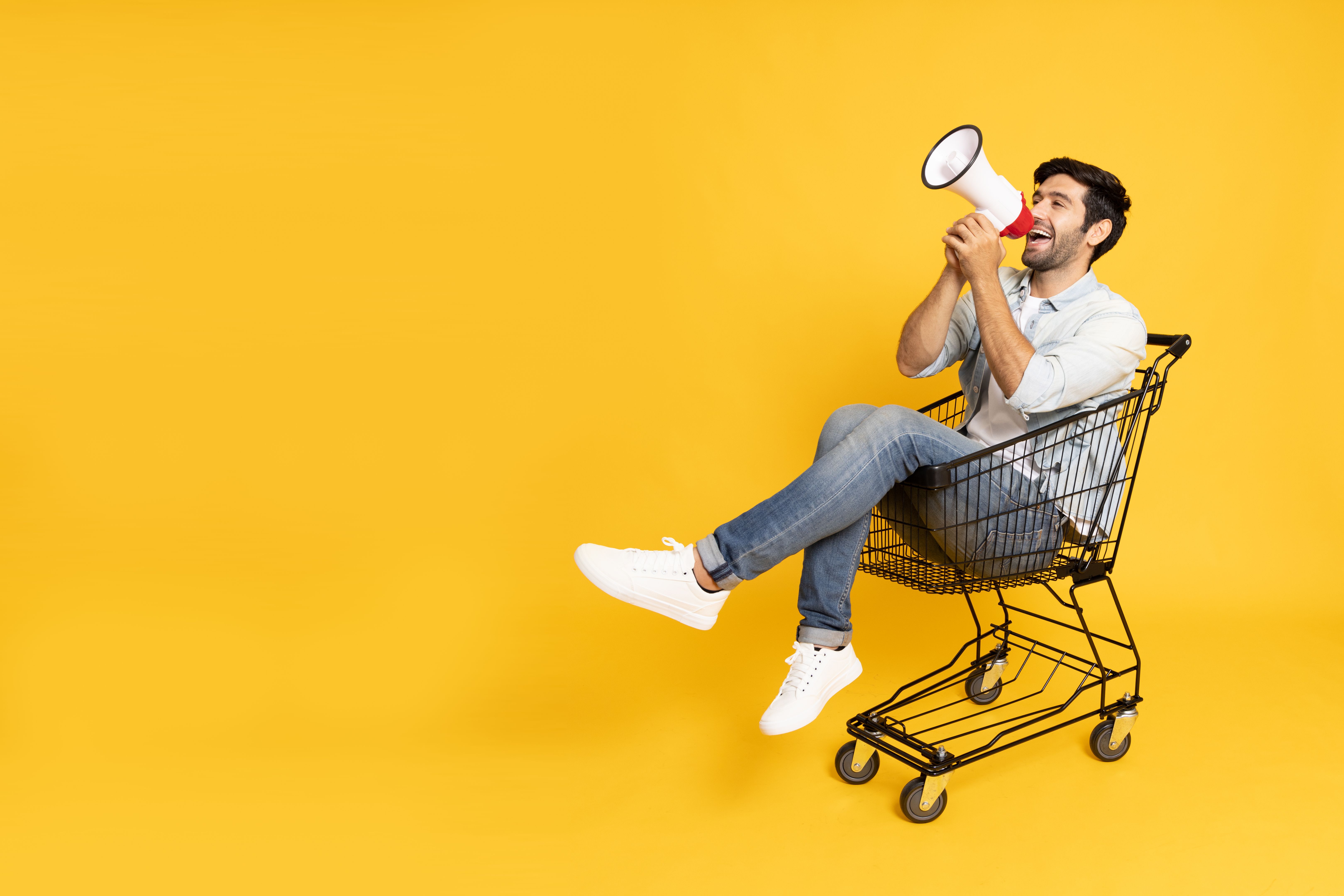Young man sitting inside of shopping trolley and holding megaphone isolated on yellow background, Announce concept